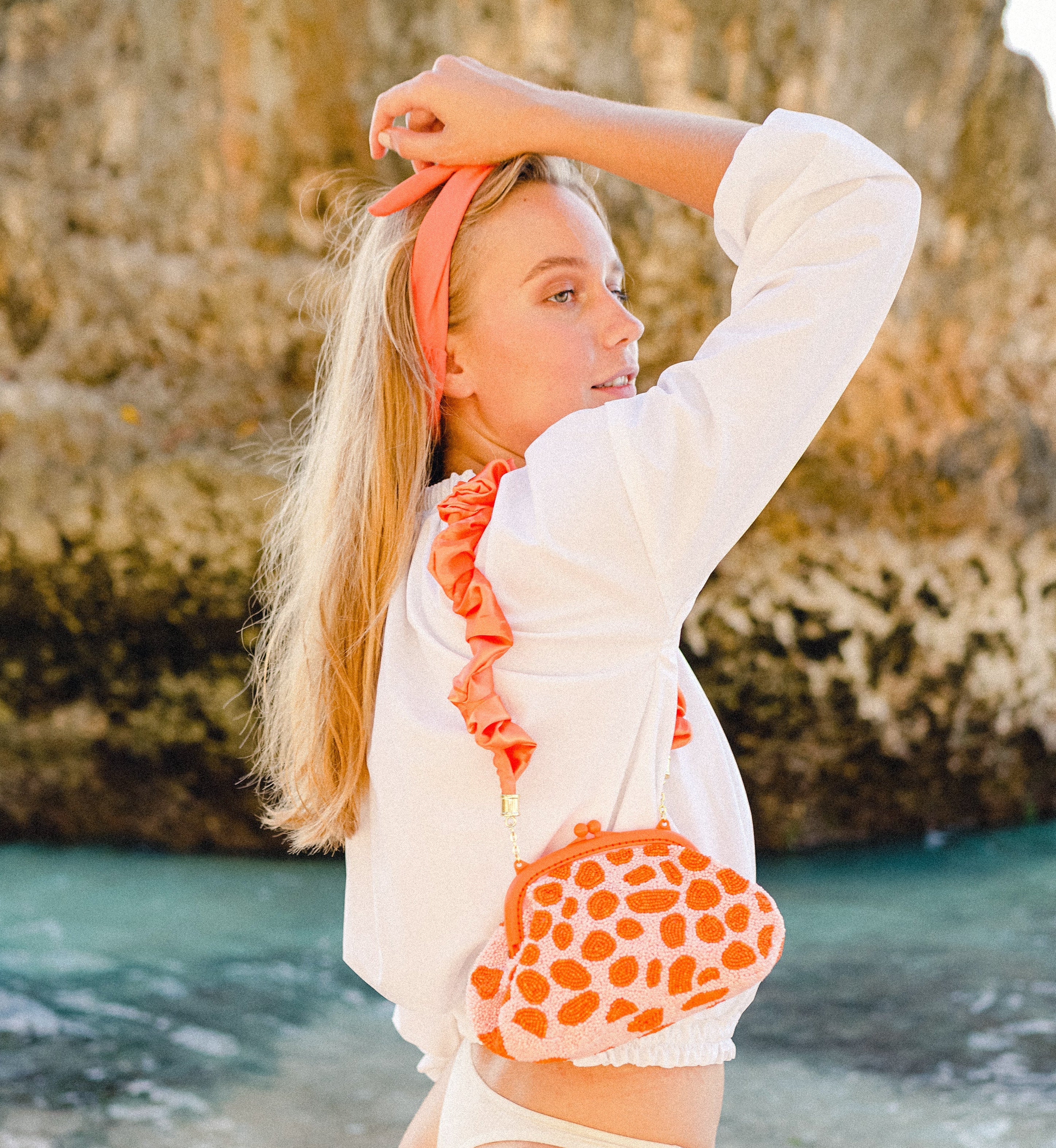 Woman on a beach wearing a white outfit with orange accents, standing in front of rocky terrain.