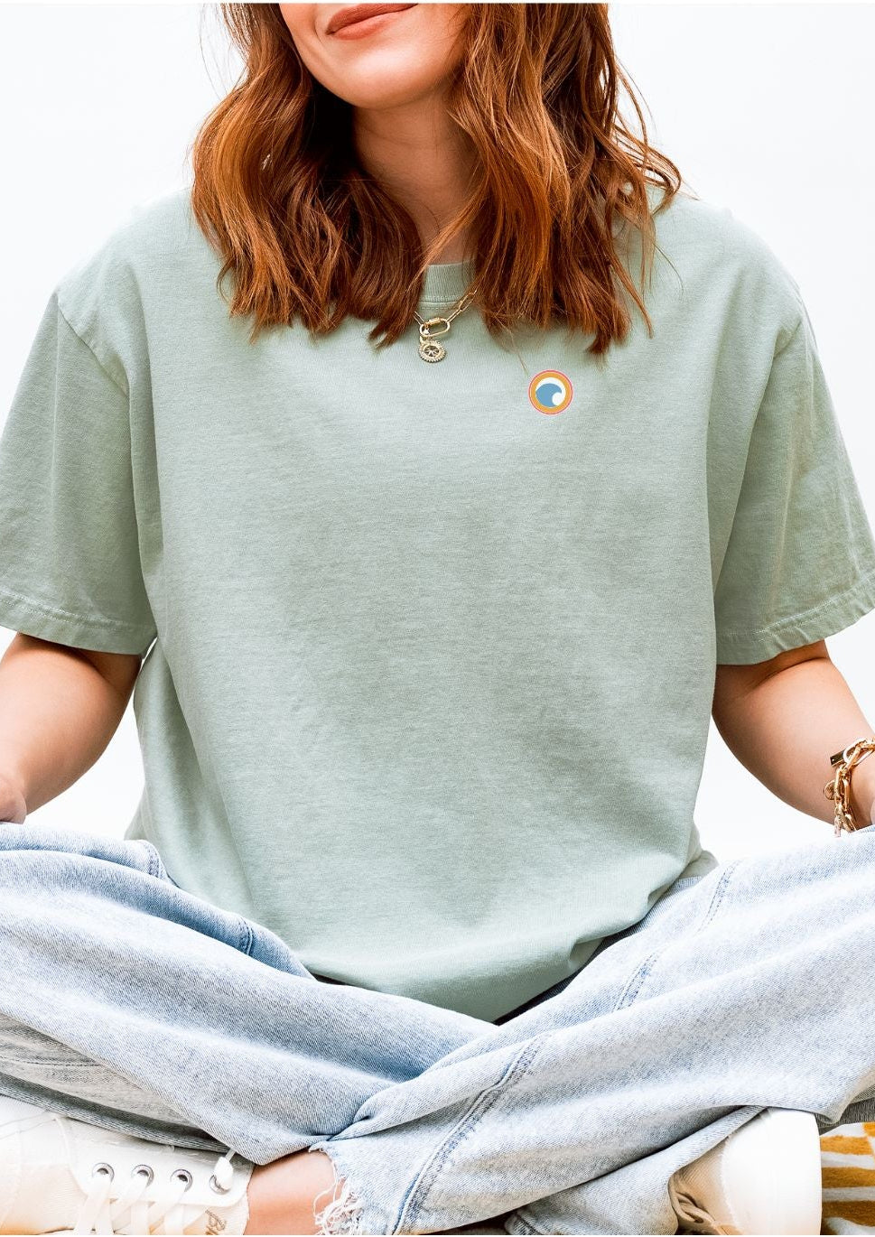 Person wearing a light green t-shirt with a logo and light blue jeans sitting on a white background.