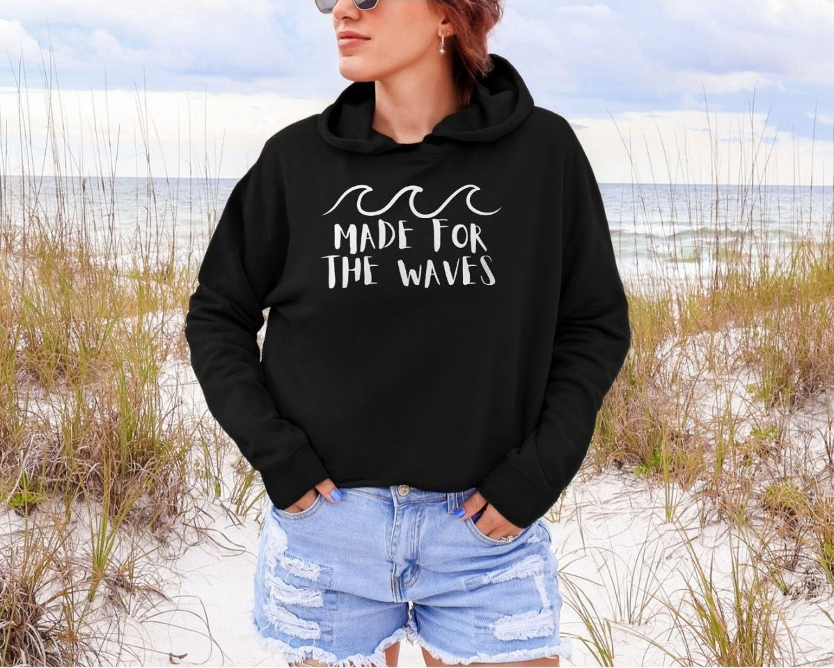 Person wearing a black hoodie with 'Made for the Waves' text on a beach.